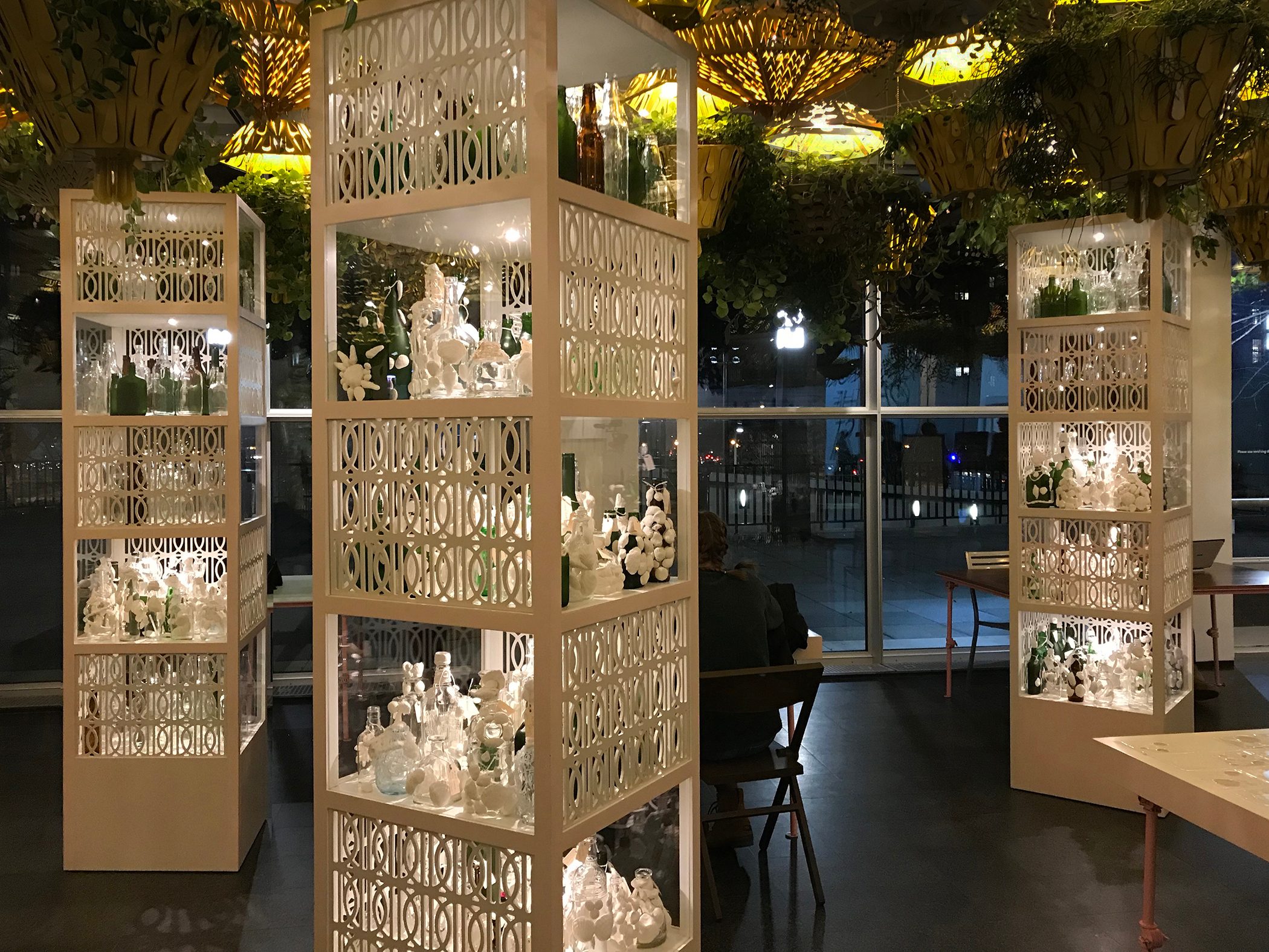 Indoor space with illuminated shelves of glass and ceramic figurines, plants overhead.