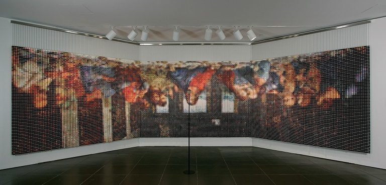 Intricate, pixelated multi-panel art installation in gallery corner under overhead lights.
