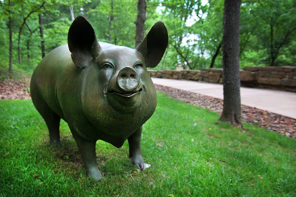 Bronze pig sculpture on grass in wooded park with stone path and trees in the background.