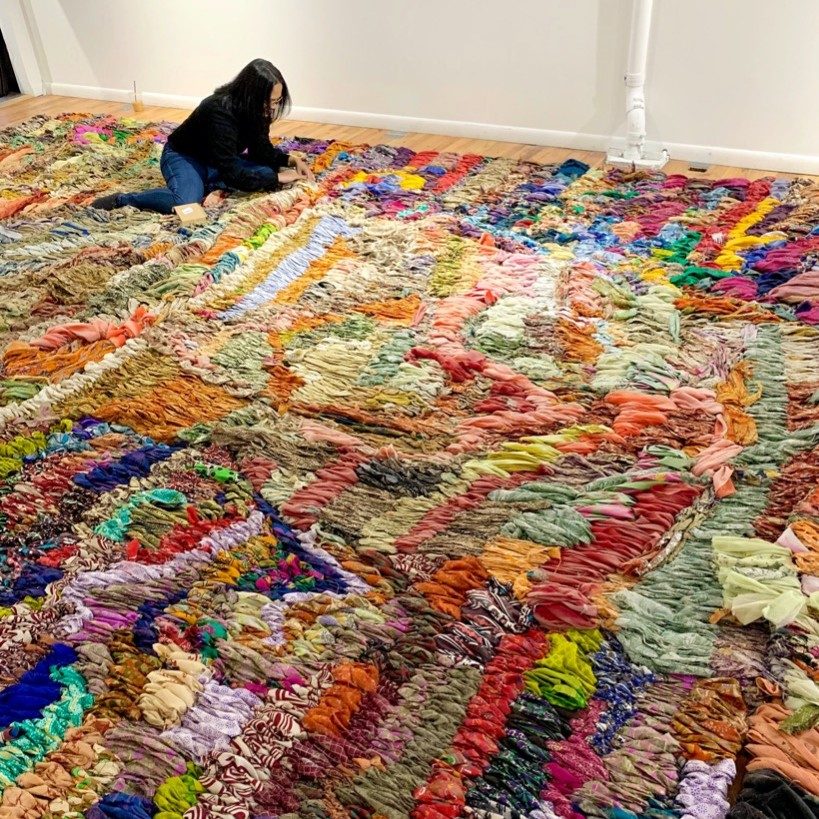 Person kneeling on a colorful textile installation indoors with fabric pieces on a wooden floor.