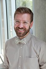 Man with glasses, beard, bow tie, smiling indoors near large windows in a long-sleeved shirt.