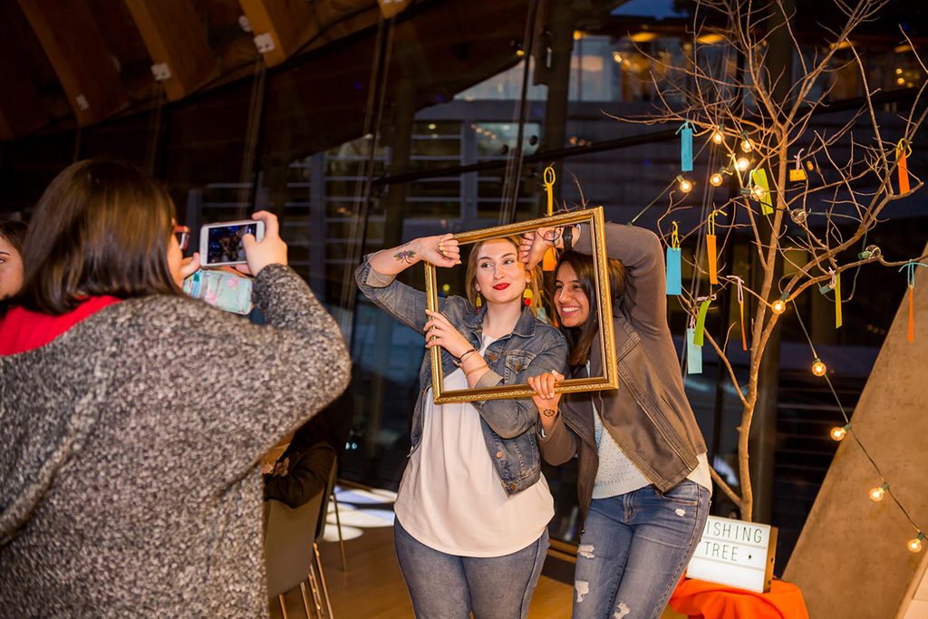 People posing with a frame for a photo near a wishing tree with colorful tags and lights.