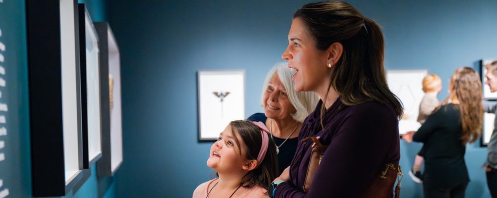 Three generations of women viewing framed exhibits on blue walls in a museum gallery.