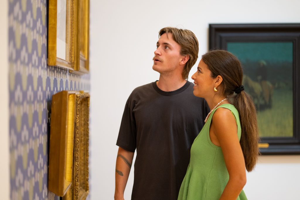 A man with a tattoo and a woman in a green top view framed paintings in a gallery.