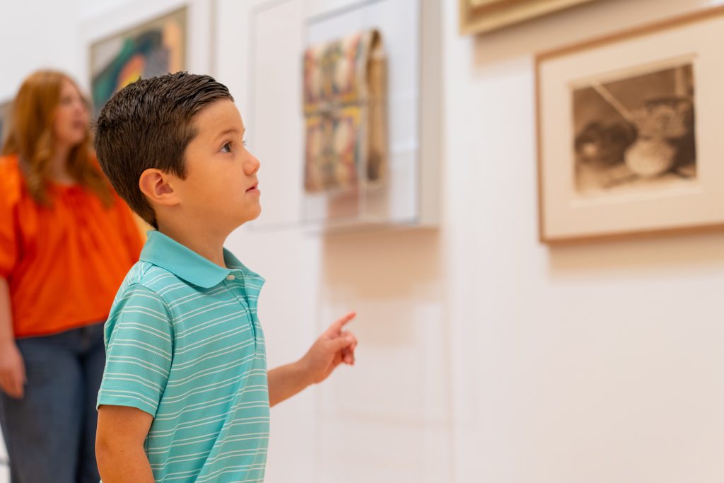 Boy in turquoise striped polo shirt viewing artwork in a gallery with a woman in the background.