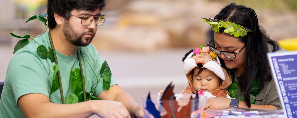 Adults with foliage accessories and child in costume crafting at blue table with art supplies.