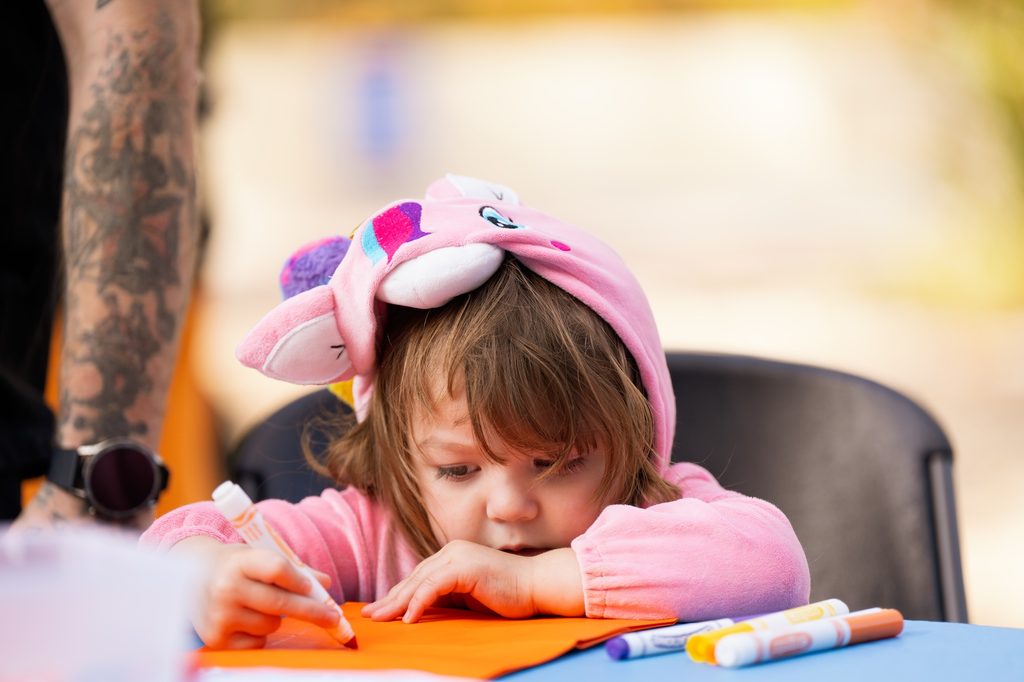 Child in pink hoodie colors on orange paper at table; adult with tattooed arm nearby.