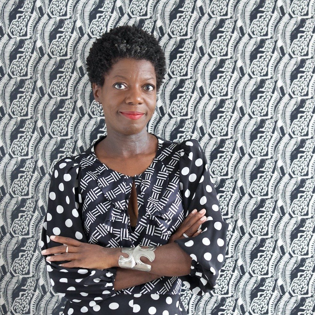 Person with short curly hair in mixed pattern dress, arms crossed, against patterned background.