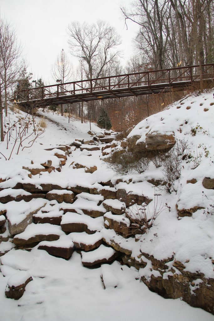 Paisaje nevado con un camino de piedra y un puente oxidado rodeado de árboles sin hojas.