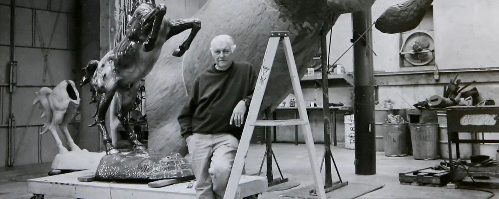 Elderly man leaning on stepladder in studio with large horse sculptures and tools.