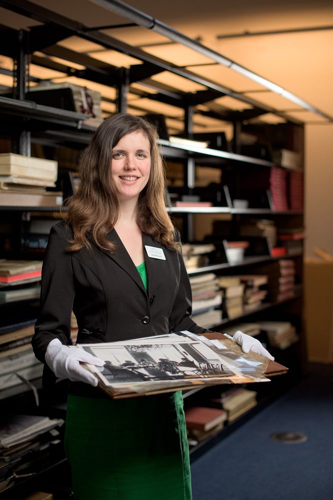 Woman in archive room holding photo, wearing black blazer, green dress, white gloves, shelves behind.