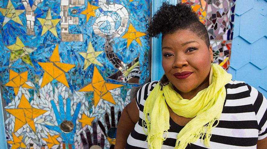 Person in striped shirt and yellow scarf by a colorful mosaic wall with stars and handprints.
