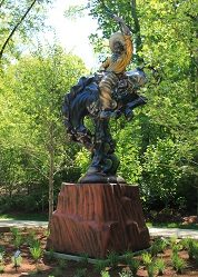 Statue of horseback rider playing guitar on wooden pedestal amid trees and greenery.