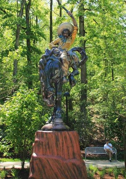 Bronze cowboy statue on rearing horse mounted on rock pedestal in a wooded park.