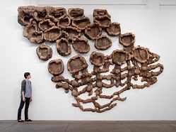 Person stands before abstract installation with wood-like, interconnected hollow structures.