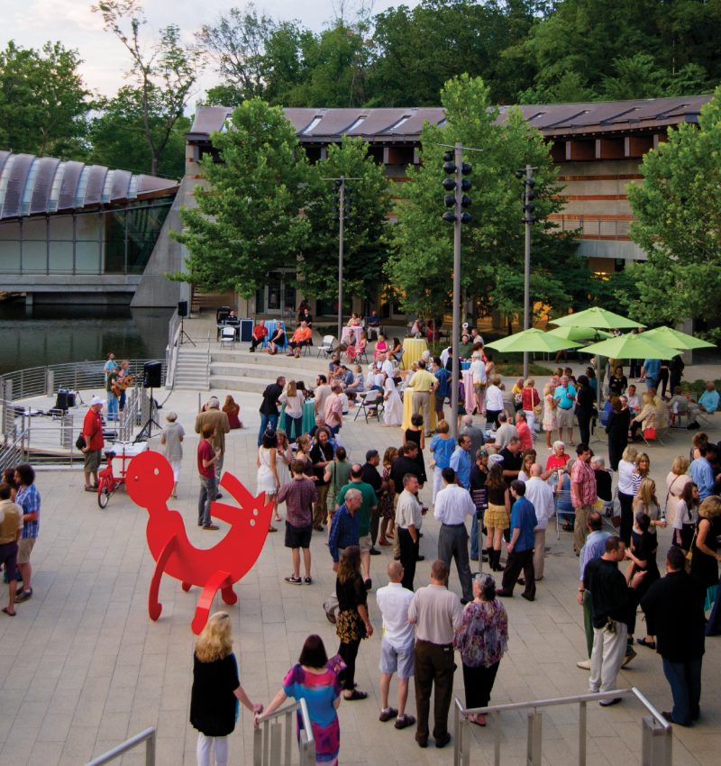 Outdoor event on patio with diverse crowd, red sculpture, tables, umbrellas, surrounded by trees.