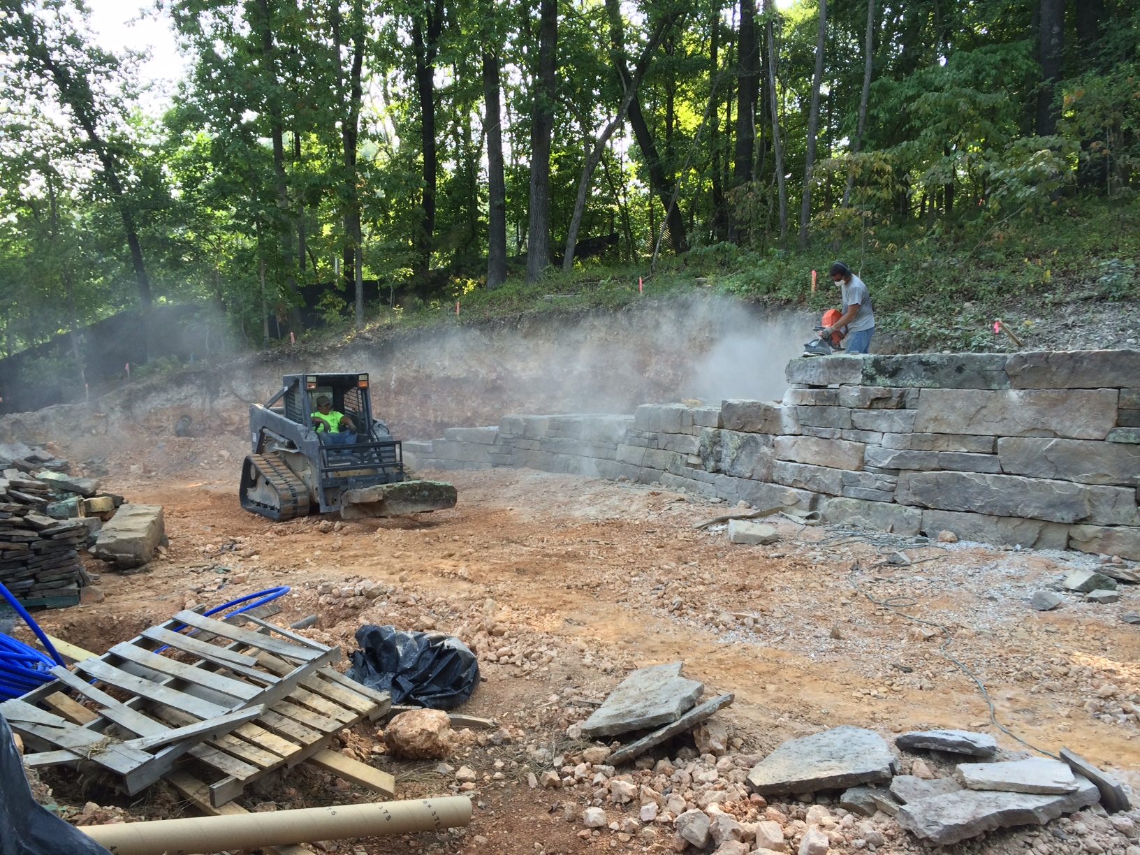 Construction site with skid-steer moving dirt, worker cutting stone for retaining wall, trees nearby.