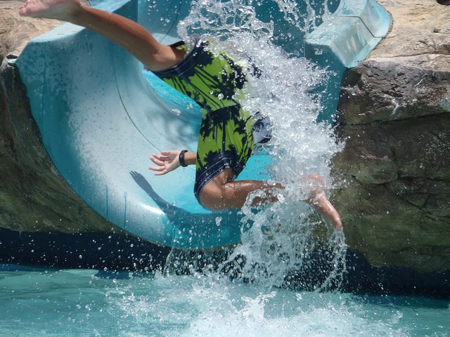 Person mid-air exiting water slide, splashing into pool, wearing swim shorts, water spraying around.