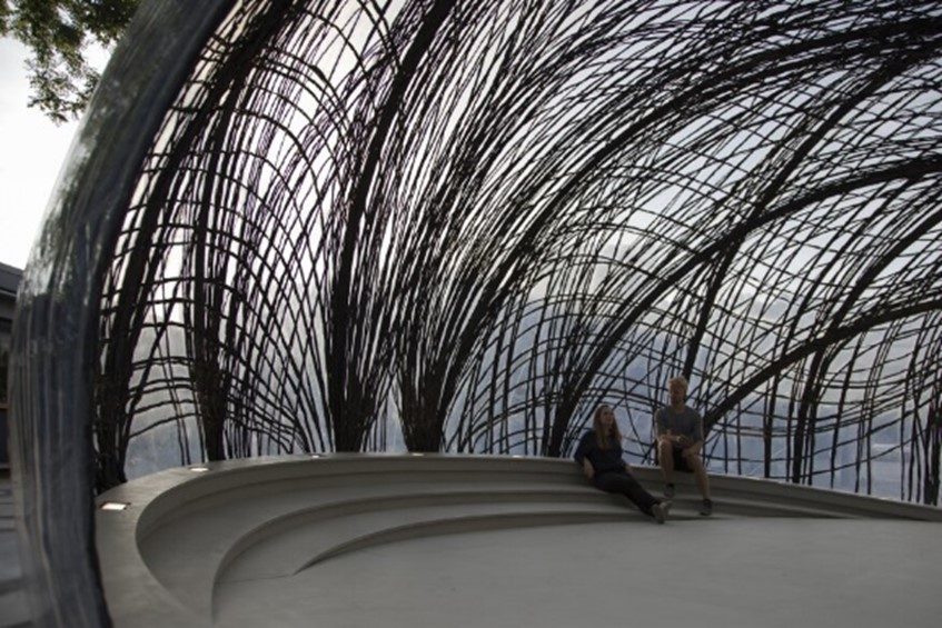 an outdoor pavilion designed in the pattern of a spiderweb with thin black metal bars intertwining to form a dome shape