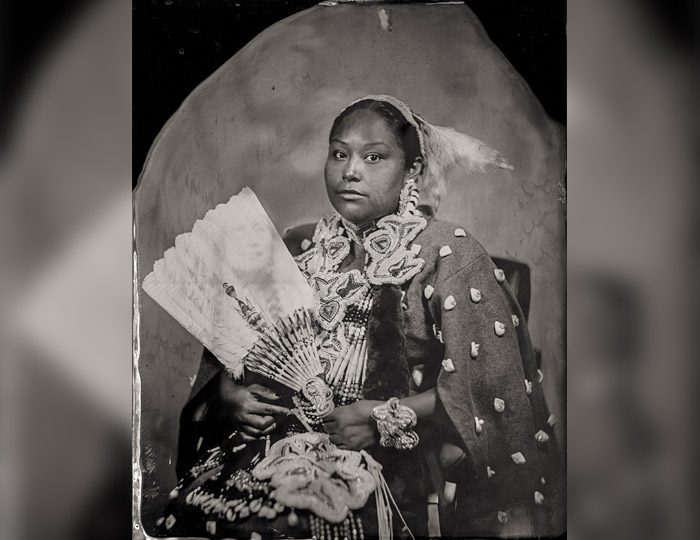 Person in intricate attire with fan, adorned with detailed designs and textures, seated for portrait.