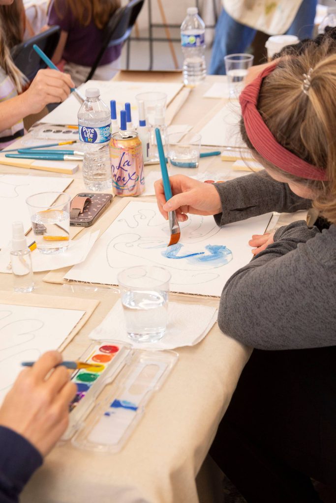 People painting with watercolors and brushes at a table with art supplies and drinks.