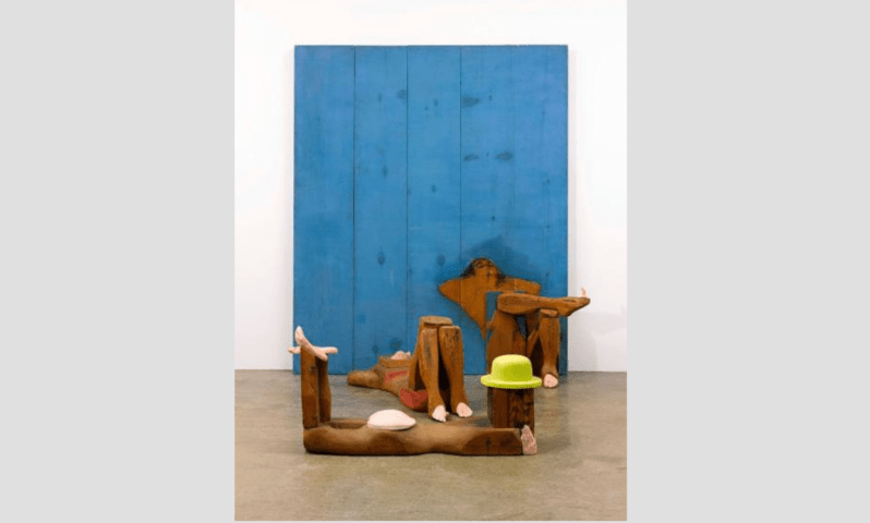 Abstract art with wooden figures, exaggerated limbs, blue backdrop, lime green hat on floor.