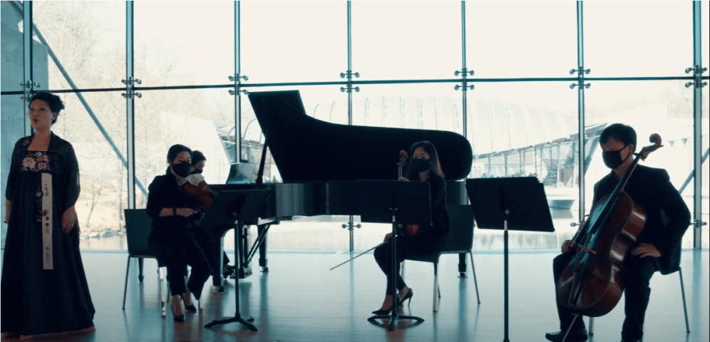 Musicians performing indoors with grand piano, violin, cello, and vocalist; formal attire, masked.