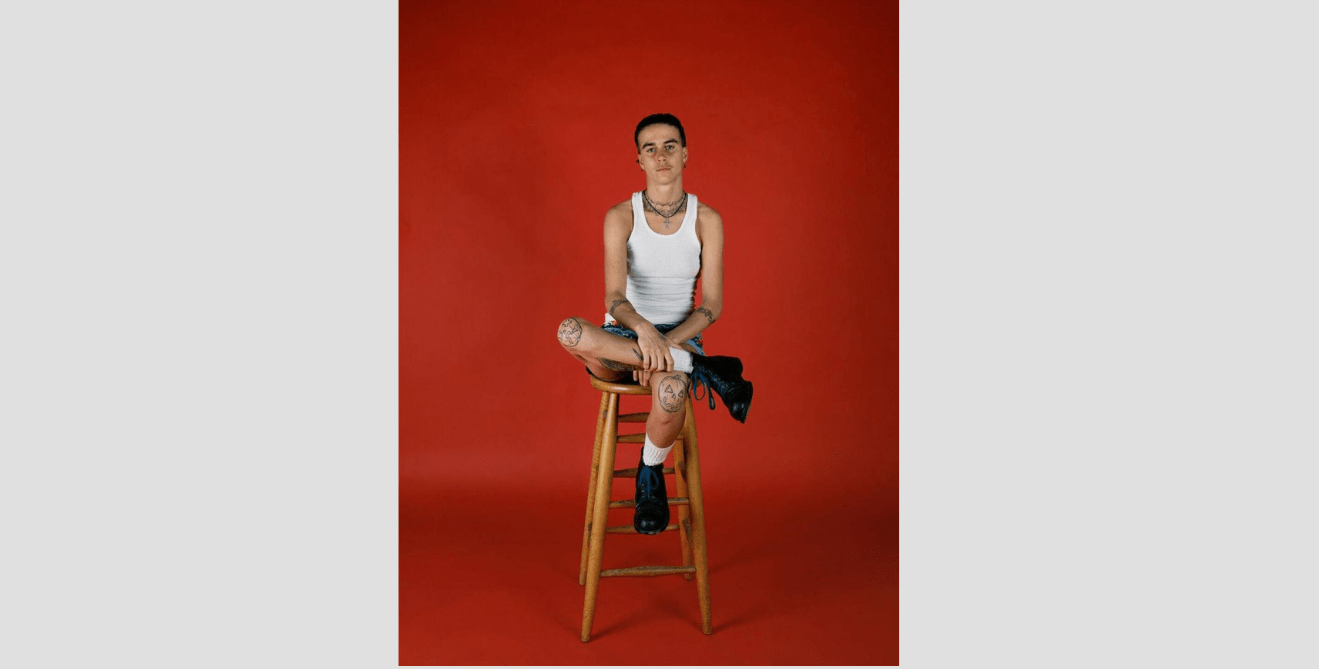 Young man in white tank shirt with tattoos on his knees sits on a stool in front of a red background