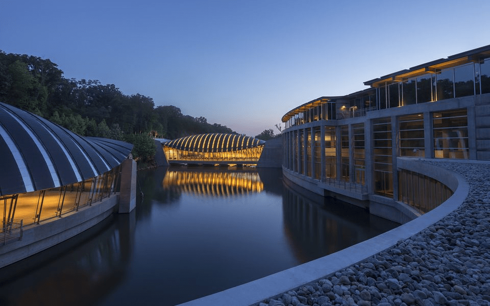 Modern buildings with curved roofs reflect on a calm waterbody at twilight, surrounded by trees.
